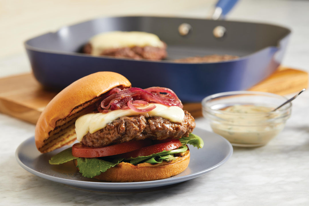 Burgers with onions and cheese on a blue plate in front of an Anolon grill pan