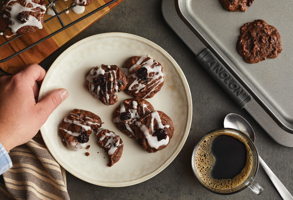 Hand holding black forest cookies on a white plate next to baking sheet, cooling rack, and coffee cup