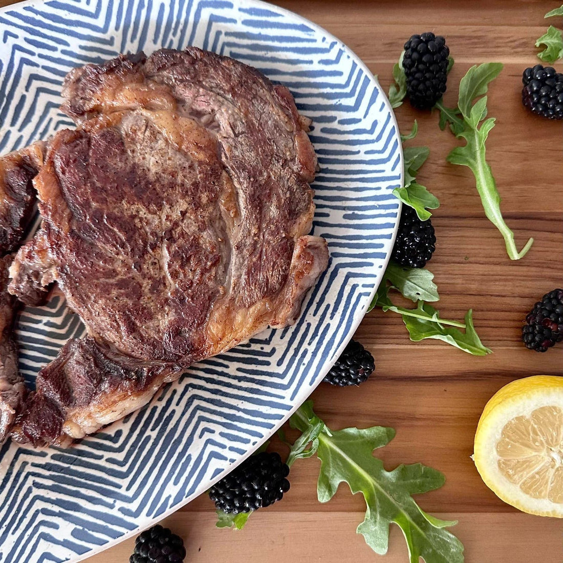 Ribeye on a blue patterned serving plate with blackberries, arugula and lemon on counter