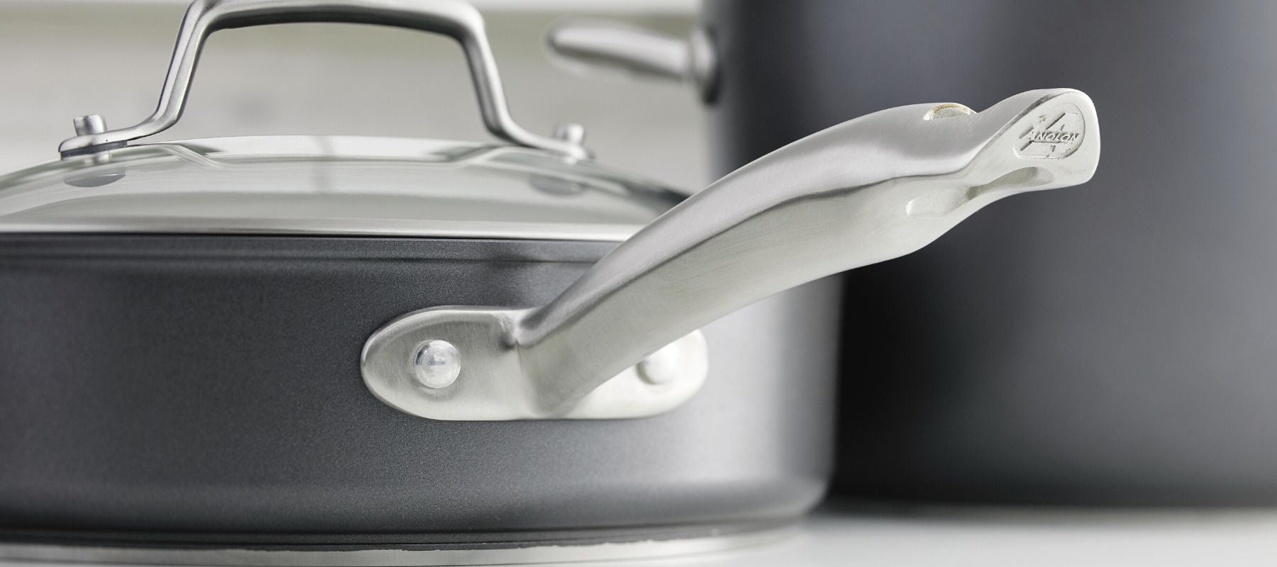 Close-up of a gray nonstick frying pan with a glass lid and a shiny stainless steel handle, placed next to a matching pot in a kitchen setting.