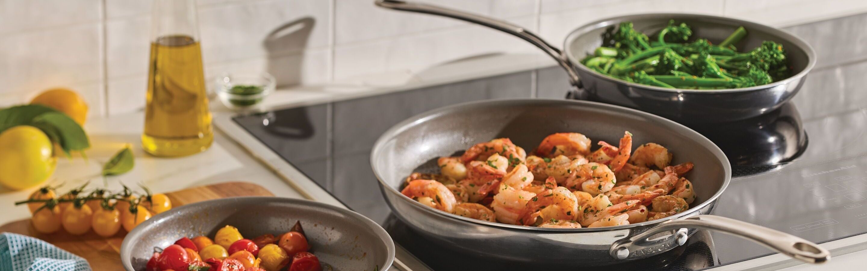 Three pans on a stovetop: one with cooked shrimp, one with cooked broccolini, and one with assorted cherry tomatoes. A bottle of olive oil and lemons are on the counter nearby.