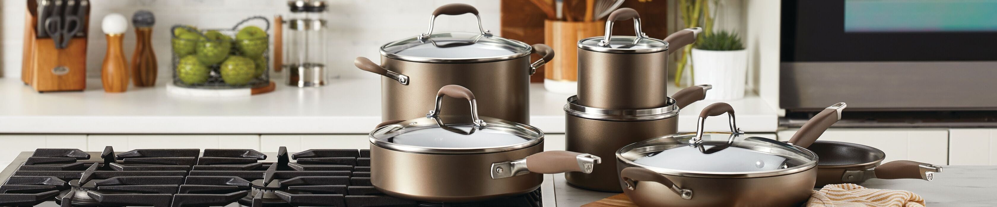 A set of pots and pans with glass lids sits on a kitchen counter and stovetop, with kitchen utensils, green apples, and a toaster visible in the background.