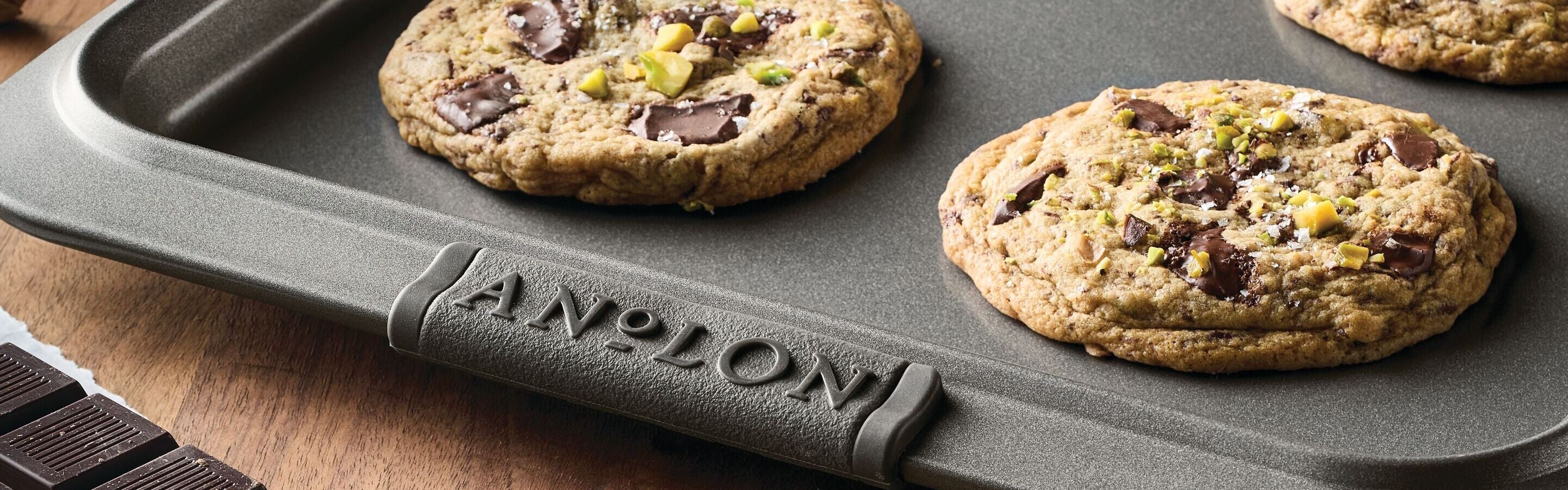 Close-up of freshly baked chocolate chip cookies with nuts on a nonstick baking tray labeled Anolon, with chocolate pieces nearby on a wooden surface.