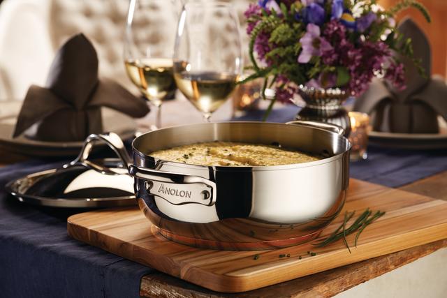 A stainless steel pot labeled Anolon filled with creamy dish on a wooden board, garnished with herbs. Set on a table with a blue cloth, wine glasses, folded napkins, and a floral centerpiece in the background.