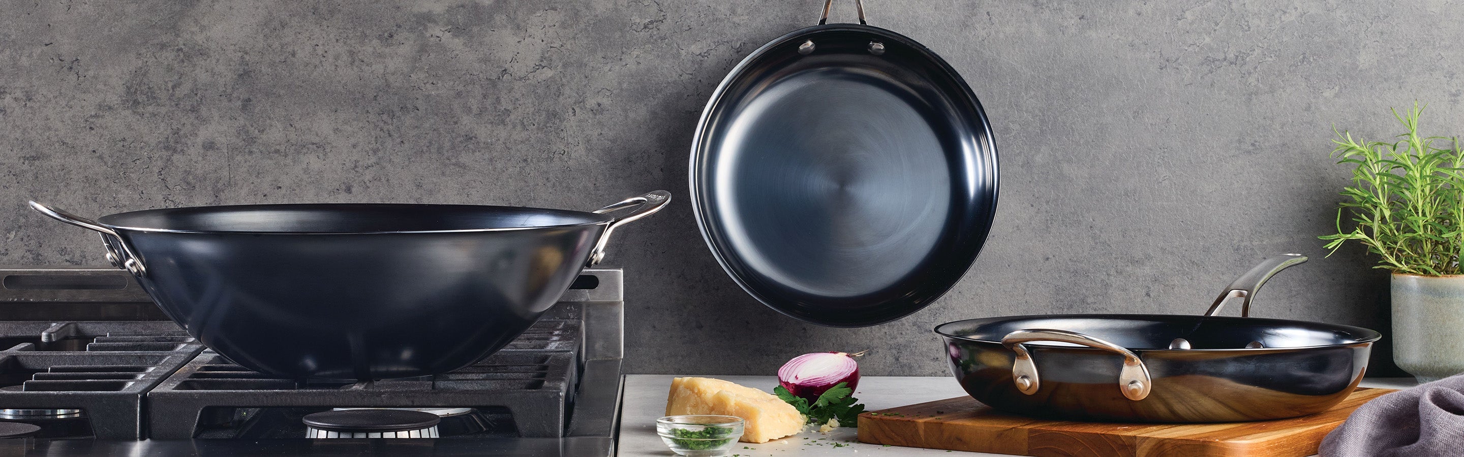 A modern kitchen scene with a wok on a stove, a frying pan hanging on the wall, and another frying pan on a wooden board beside herbs, cheese, and a red onion, against a gray background.
