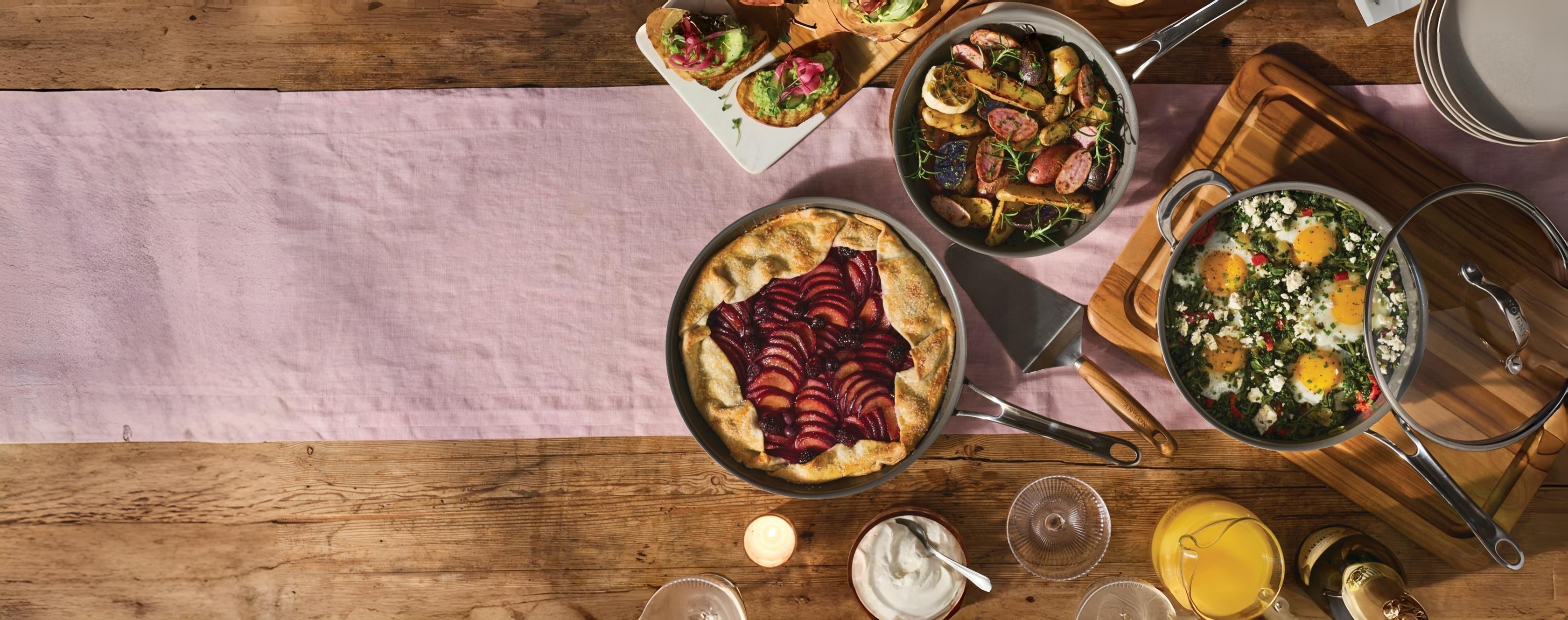 Tablescape featuring a variety of prepared dishes in Anolon cookware set on a wooden table with a pink linen runner.