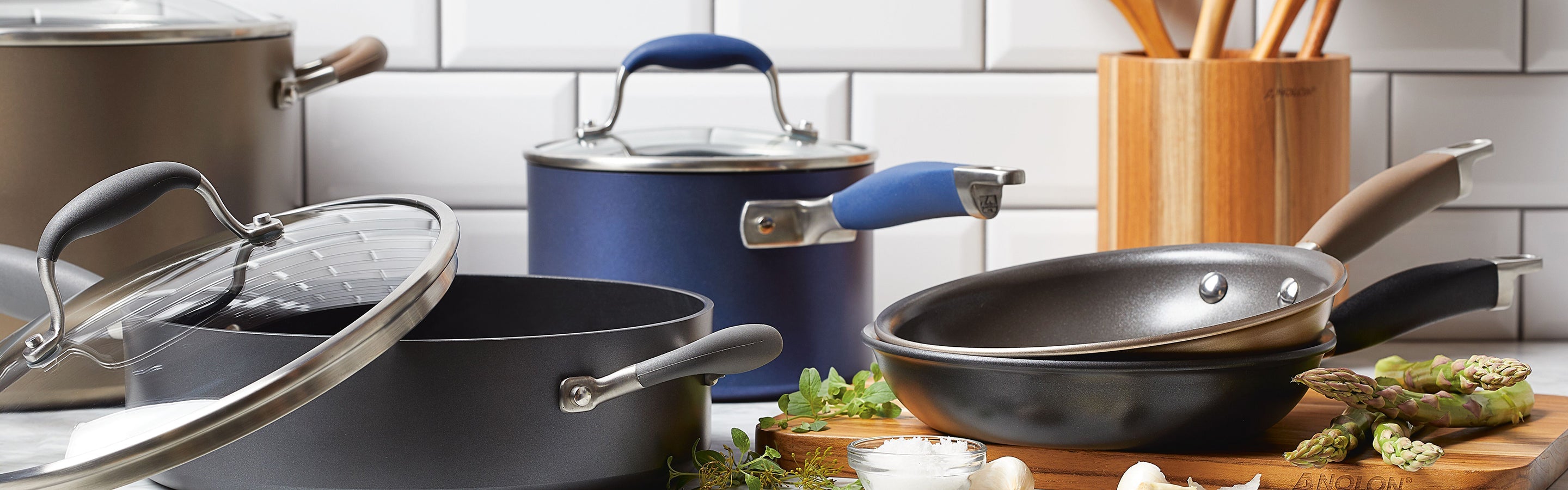 An assortment of nonstick pots and pans with lids, utensils in a wooden holder, garlic, asparagus, and herbs on a kitchen counter with a white tile backsplash.