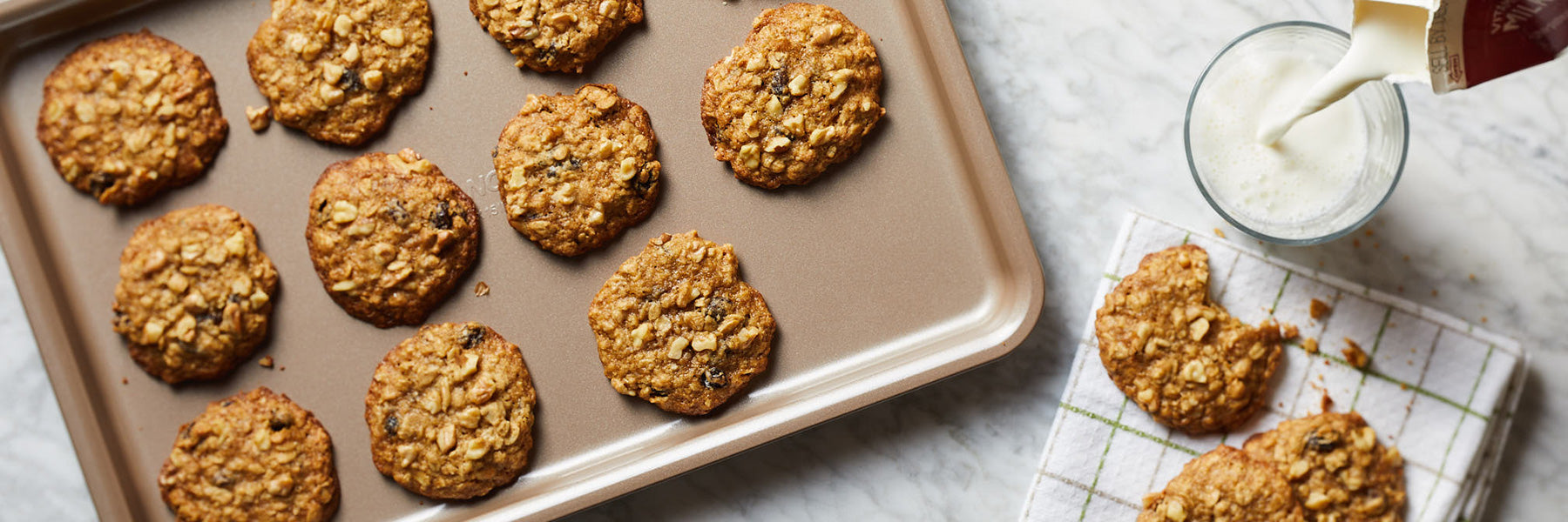 A tray of freshly baked oatmeal cookies on a marble surface, with some cookies on a napkin next to a glass of milk being poured.