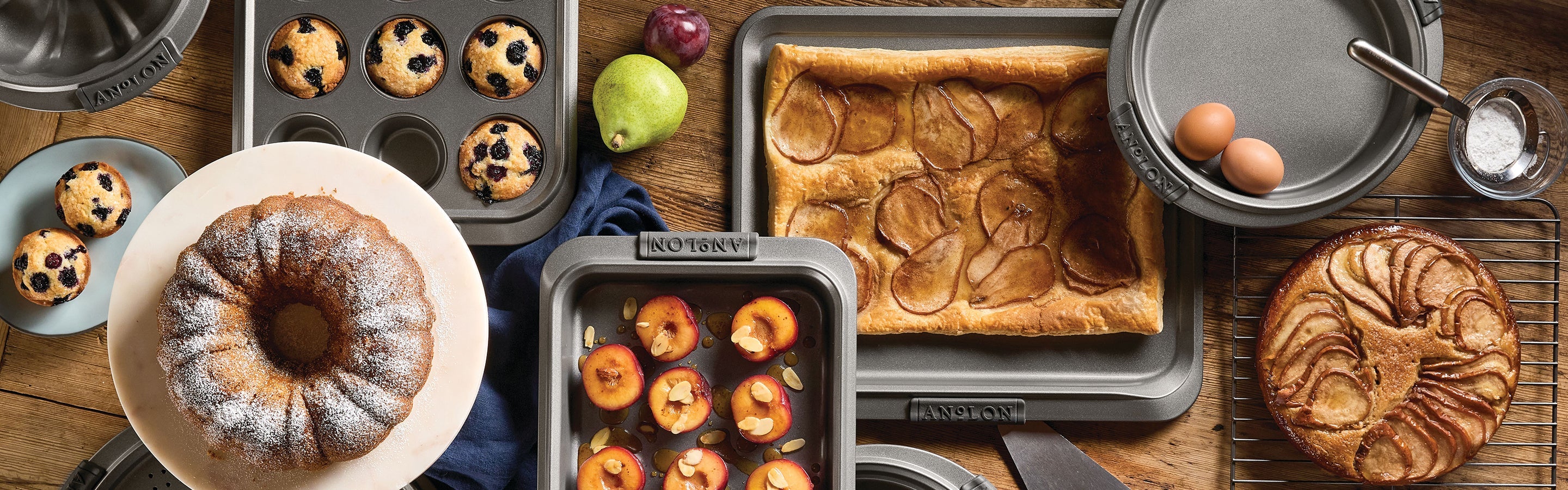 Assorted baked goods including pastries with tomatoes and basil, muffins with chives, and a bread pudding in rectangular and muffin tins on a wooden surface, with scattered blueberries.