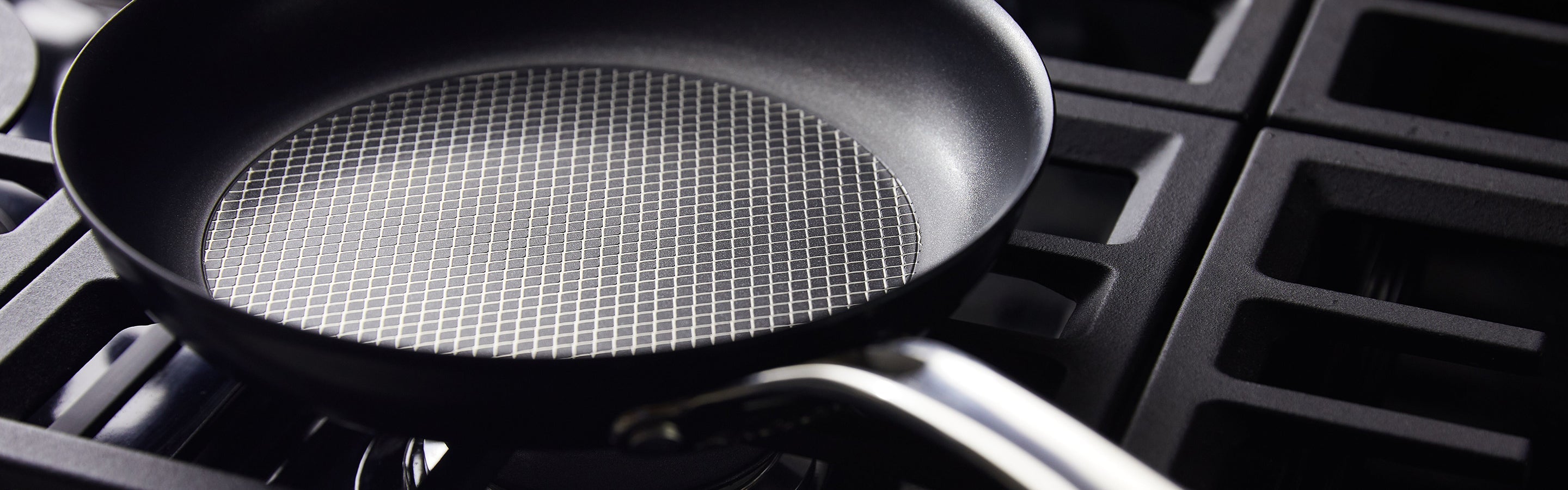 A set of black non-stick pots and pans with silver handles placed on a wooden countertop. A few onion skins are scattered nearby, with a folded cloth on the left. The backdrop has a geometric pattern.