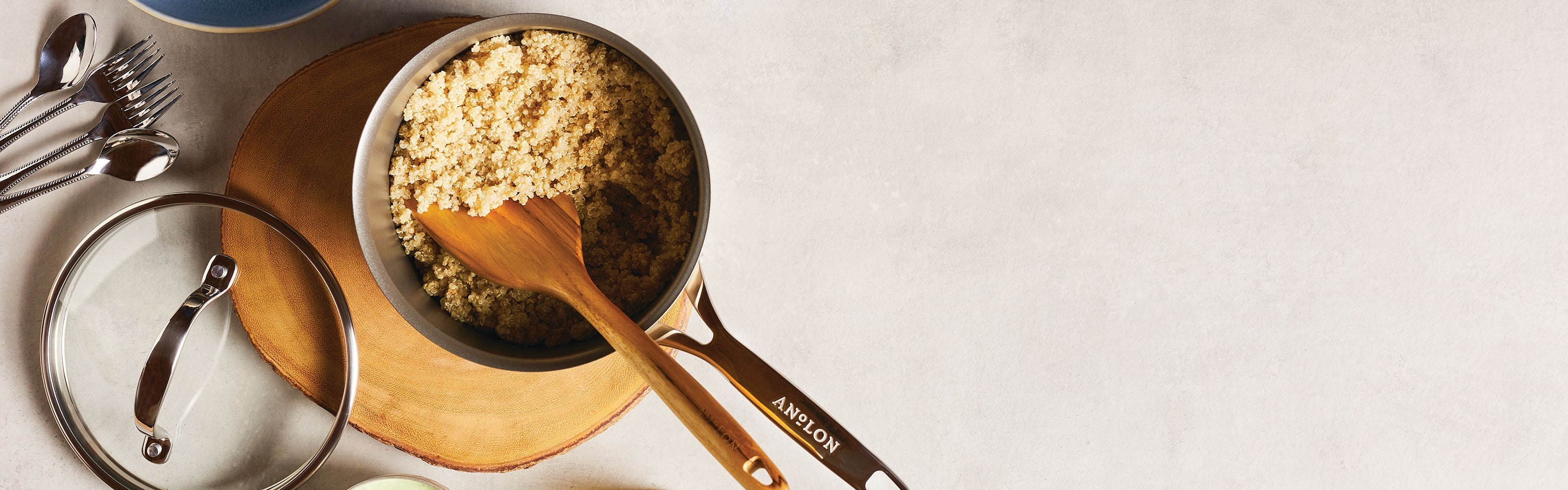 A saucepan filled with cooked quinoa sits on a wooden trivet, with a wooden spoon resting inside. A lid and some silverware are placed nearby on a light-colored surface.