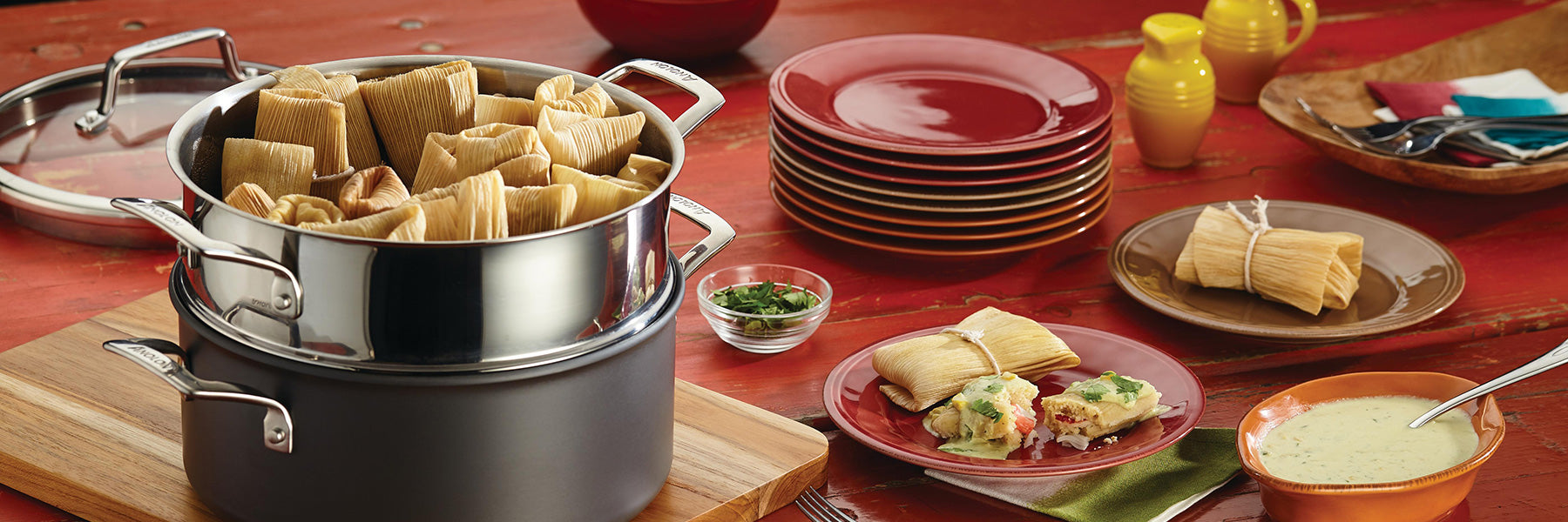 A steamer pot filled with tamales sits on a wooden board. Nearby are plates, utensils, tamales tied in corn husks, a small bowl of sauce, and dishes on a red table.