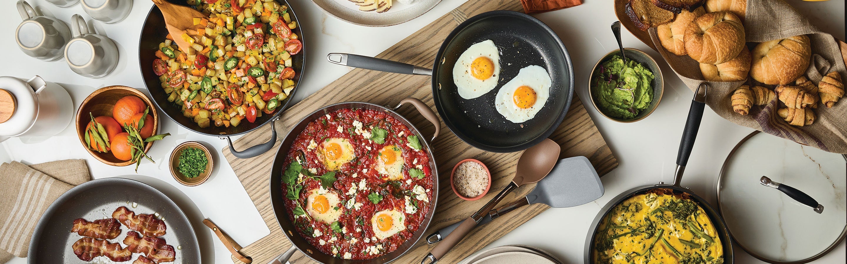 A breakfast spread with fried eggs, shakshuka, sautéed vegetables, bacon, an omelette, bread rolls, avocado, fresh tomatoes, herbs, cups, and plates on a white table.