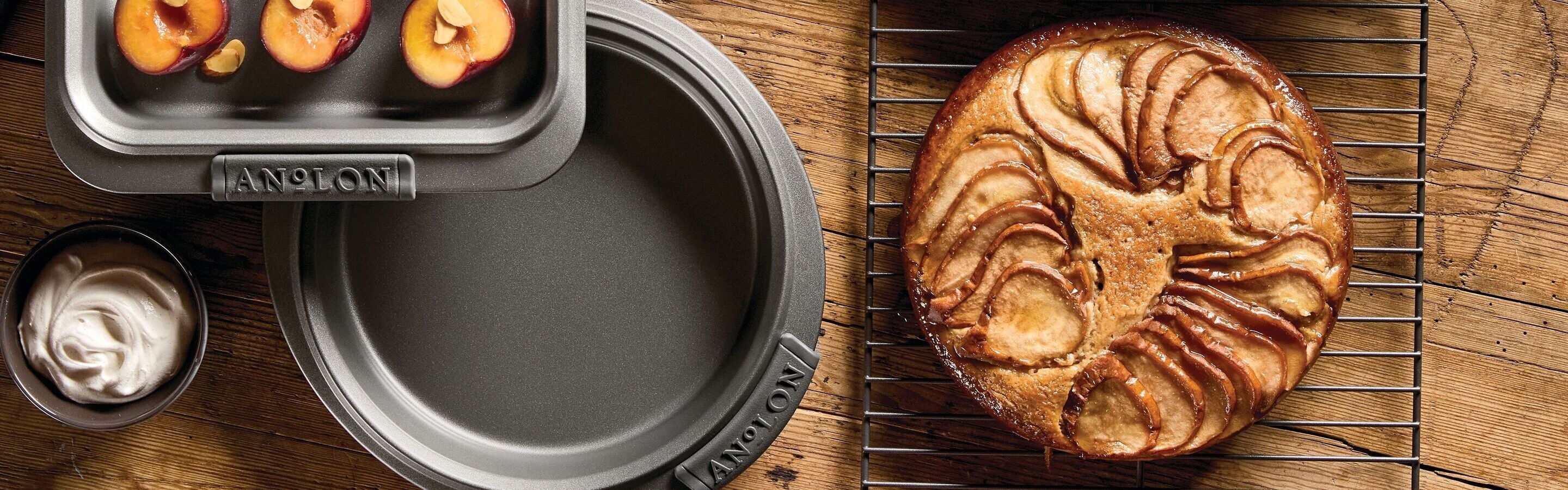 Baked apple dessert on a cooling rack, with empty baking pans, roasted apples in a pan, and a bowl of whipped cream on a wooden table.