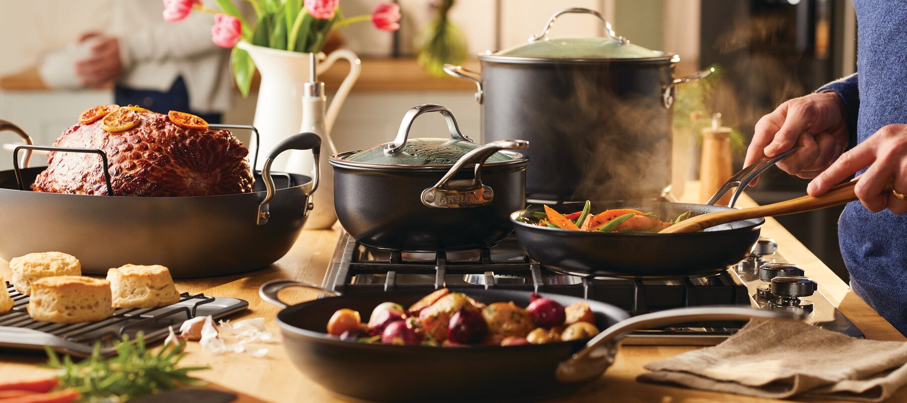 A person cooks vegetables in a skillet on a stove, surrounded by pots, a roasted ham, biscuits, and a plate of mixed vegetables in a cozy kitchen setting.