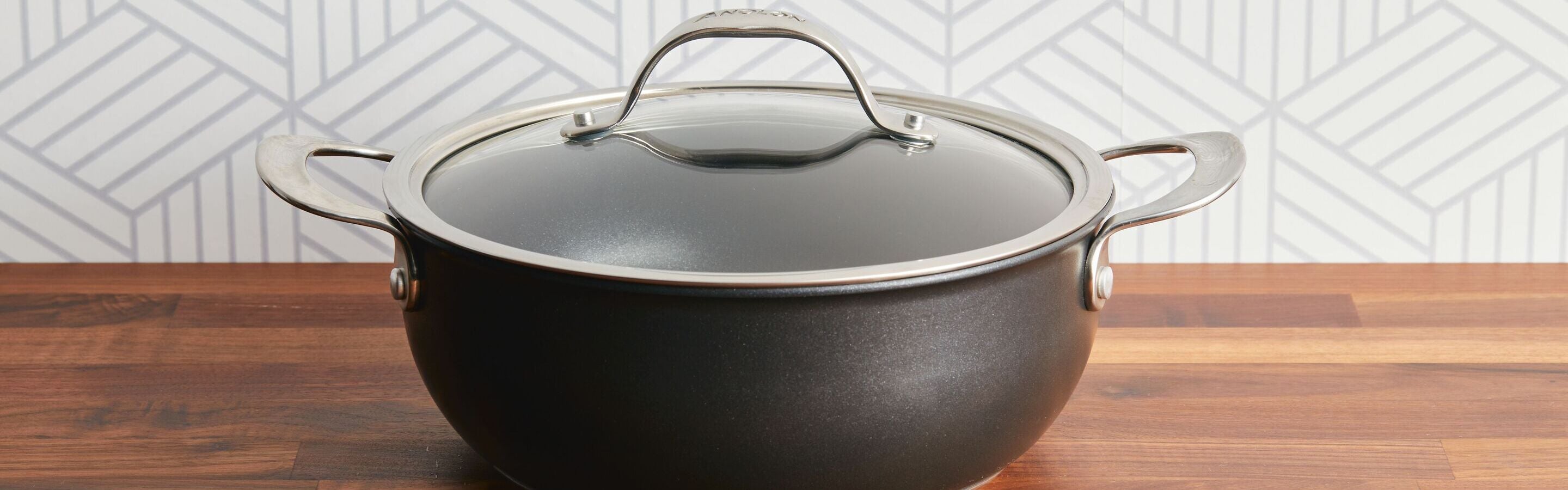 A black cooking pot with a glass lid and metal handles sits on a wooden countertop against a geometric patterned wall.