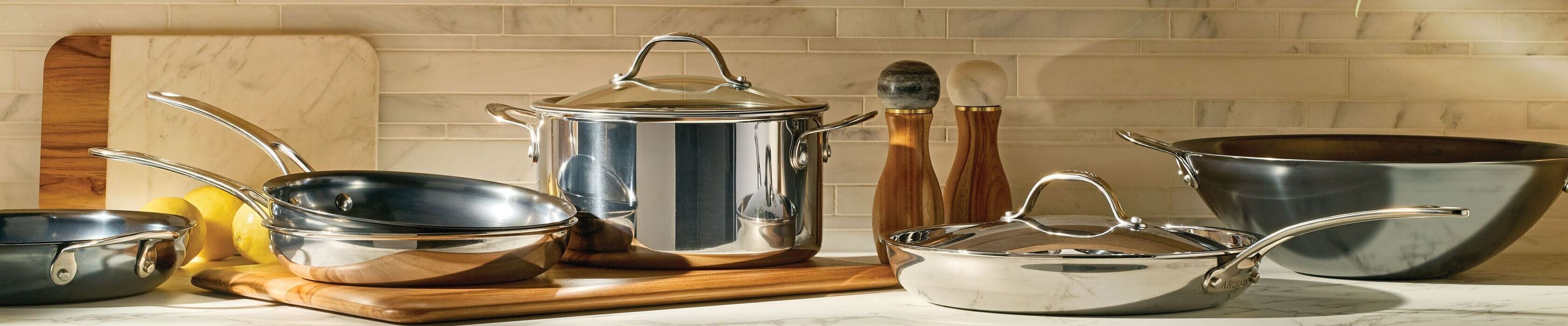 Three stainless steel and nonstick pans on a marble countertop with a wooden cutting board, wooden spoon, spatula, and green leaves in sunlight.