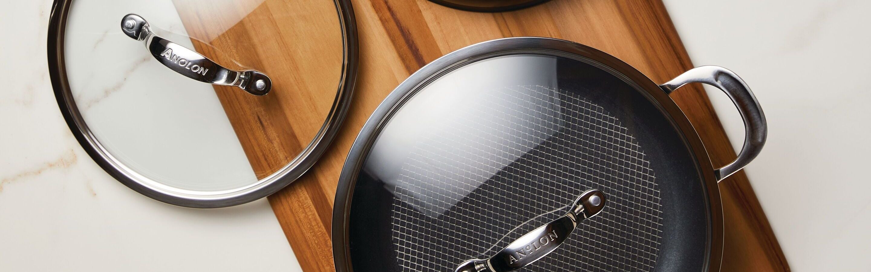 Two glass pot lids and a nonstick frying pan with a glass lid are arranged on a wooden cutting board on a white surface. The cookware features metal handles.