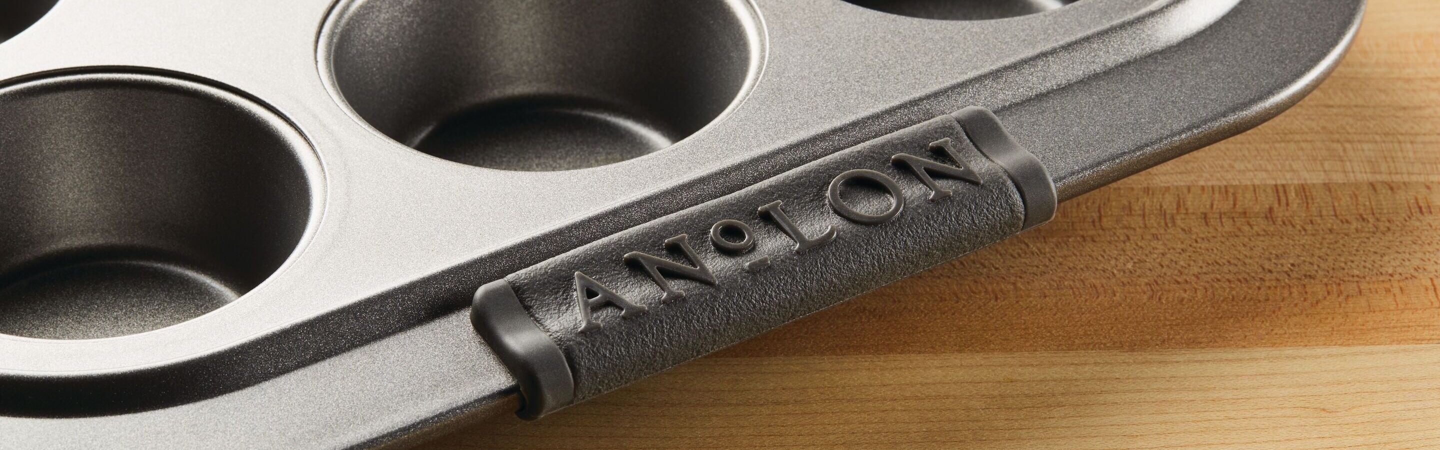 A metal muffin pan with the brand name ANOLON embossed on its handle sits on a wooden surface.