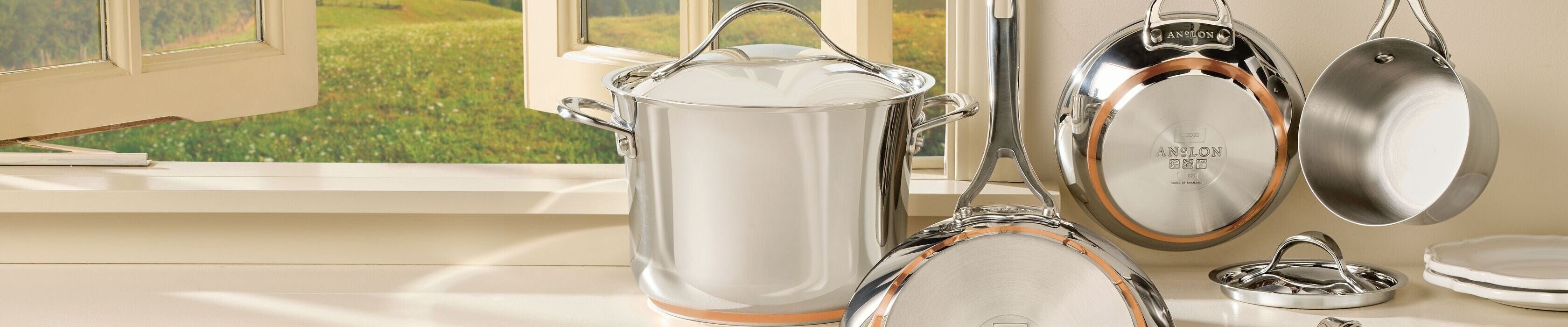 A set of shiny stainless steel pots and pans with copper accents on a kitchen counter and hanging on a rack, with a window showing a sunny outdoor landscape in the background.