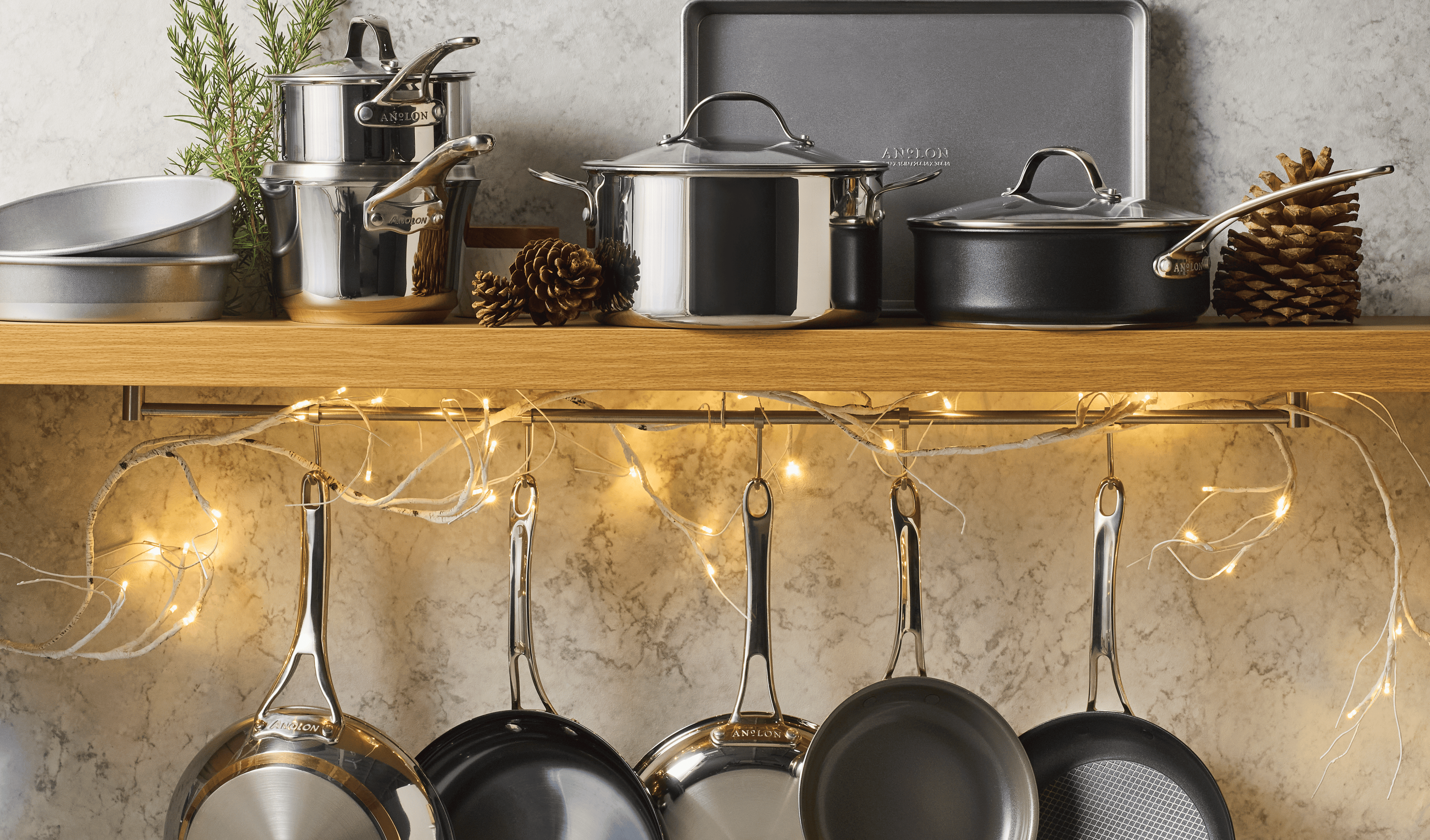 A wooden kitchen shelf holds pots, pans, and baking trays, decorated with pinecones, rosemary, and string lights. More frying pans hang from hooks below the shelf against a marble backsplash.