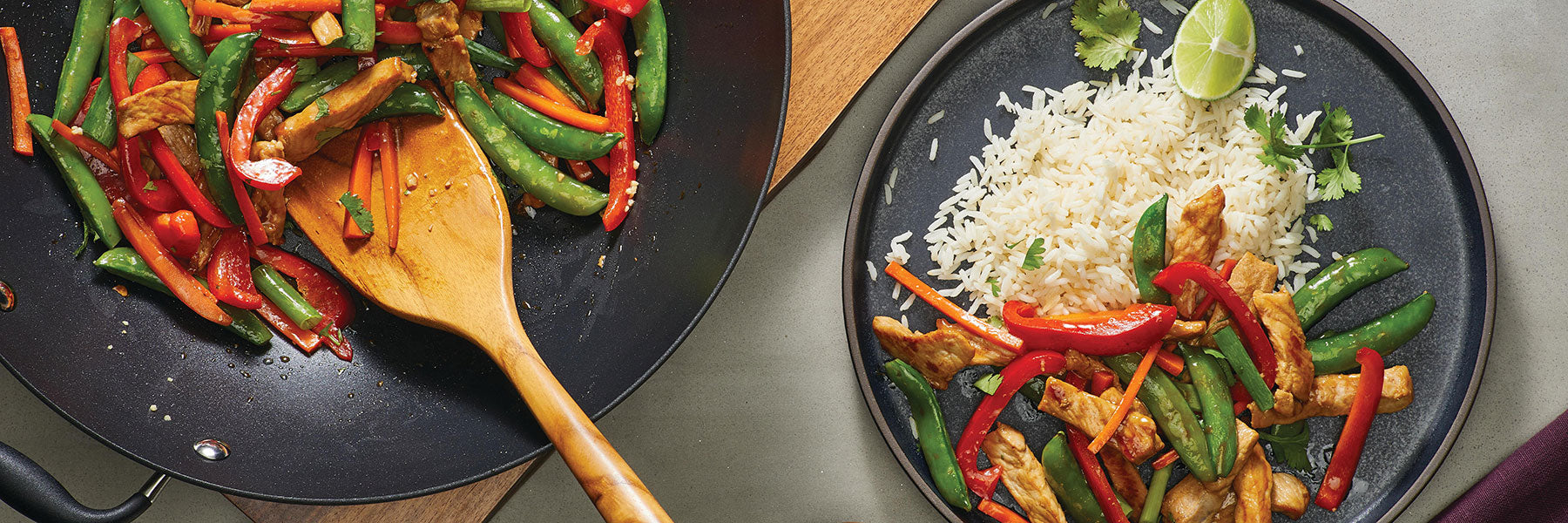 A pan of colorful stir-fried vegetables and chicken is next to a plate with the stir-fry served alongside white rice, garnished with cilantro and a lime wedge.
