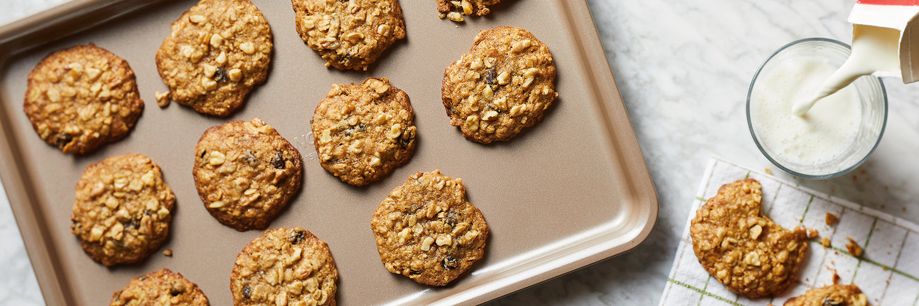 A baking tray with freshly baked oatmeal raisin cookies on a marble counter. A glass of milk is being poured on the right, next to a plate of cookies.