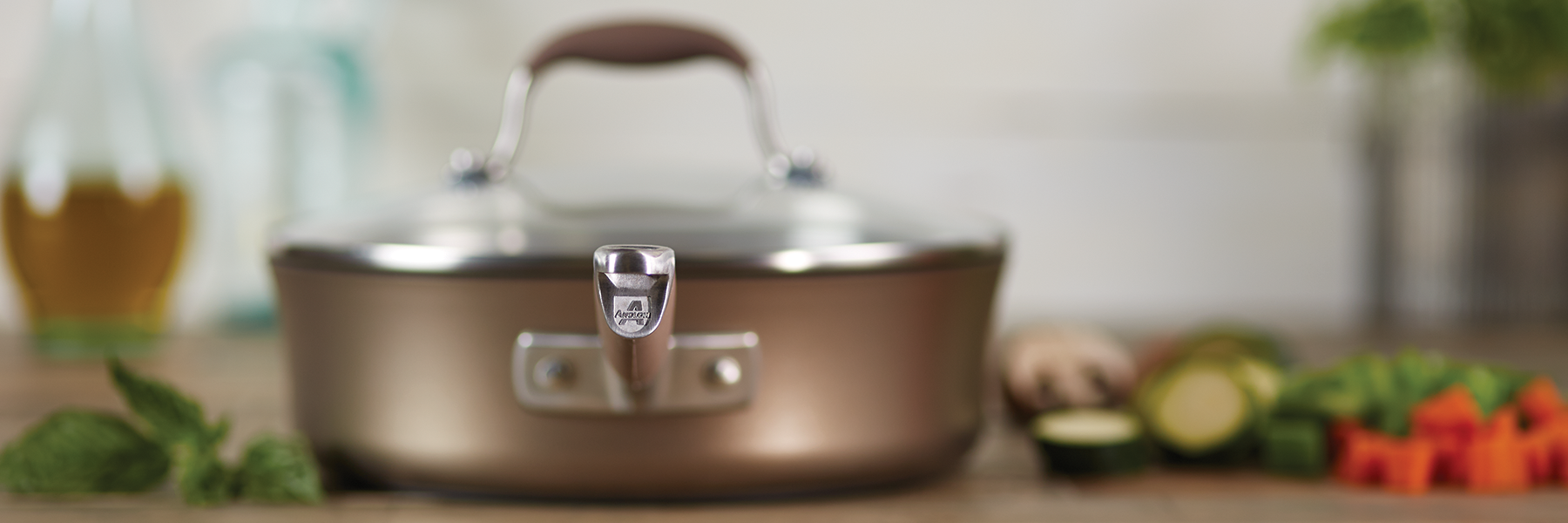 A stainless steel pan with a lid sits on a kitchen counter, surrounded by fresh vegetables, herbs, and bottles in the blurred background.