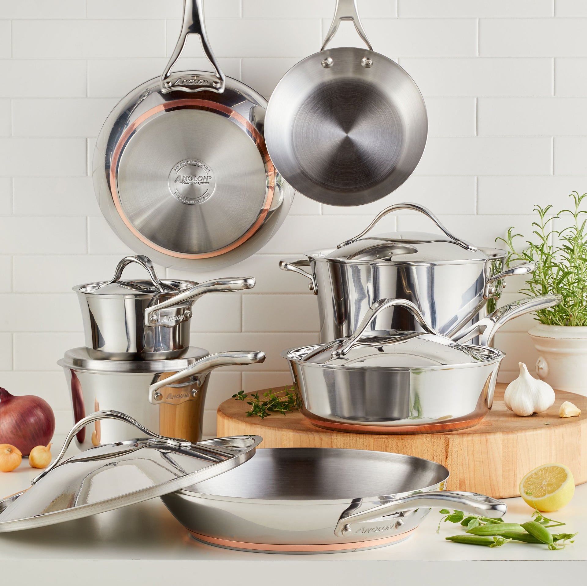 A professional lifestyle photograph of the Anolon Nouvelle Copper Stainless Steel 12-piece cookware set. The collection is arranged on a light wooden butcher block and features polished stainless steel exteriors with distinct copper mid-layers at the base. The set includes various sized saucepans, a stockpot, and frying pans, some hanging from a pot rack against a white subway tile backsplash, surrounded by fresh ingredients like lemon, garlic, and snap peas.