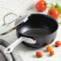 The Anolon X 2.5-Quart Hybrid Nonstick Saucier Pan with a stainless steel handle and glass lid sits on a marble countertop. Cherry tomatoes and regular tomatoes, accented by green leaves, rest nearby against a geometric-tile backsplash. Its safe for metal utensils thanks to its durable SearTech™ technology.
