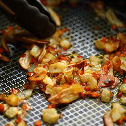 A close-up of golden, caramelized chopped vegetables like potatoes and red peppers roasted in an Anolon X 3.5-Quart Hybrid Nonstick Induction Sauté Pan with Lid, stirred by tongs on a grid-patterned basket using SearTech technology.