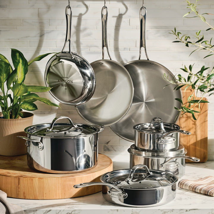 An EverLast Tri-Ply Clad Stainless Steel 11-Piece Cookware Set with lids sits on a kitchen counter, surrounded by wooden boards, green potted plants, and set against a white tile backsplash.