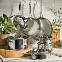 An EverLast Tri-Ply Clad Stainless Steel 11-Piece Cookware Set with lids sits on a kitchen counter, surrounded by wooden boards, green potted plants, and set against a white tile backsplash.
