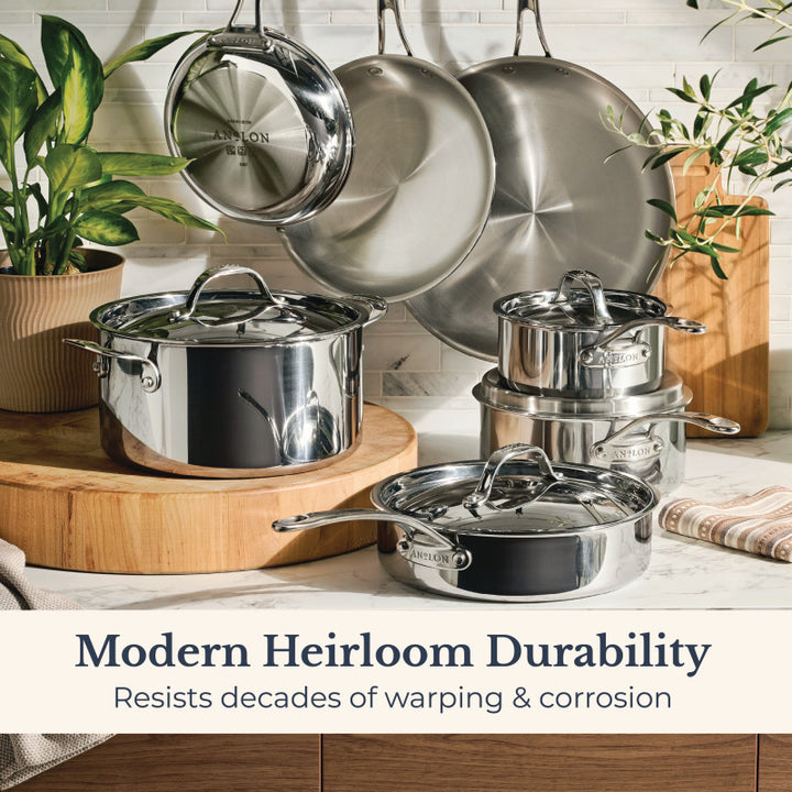 An 11-Piece EverLast Tri-Ply Clad Stainless Steel Cookware Set with lids sits on a kitchen counter and hangs on the wall among plants. Text: “Modern Heirloom Durability—Resists decades of warping & corrosion.”.