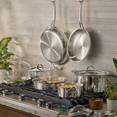 A modern kitchen stove features the EverLast Tri-Ply Clad Stainless Steel 11-Piece Cookware Set in use, with two pans hanging on the backsplash and green plants decorating the counter.