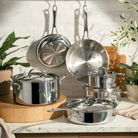 A lifestyle shot of the Anolon EverLast Tri-Ply Clad stainless steel 10-piece cookware set, featuring several pots and pans with lids arranged on a marble countertop and two frying pans hanging from a rack against a white tiled wall, accented by green plants and wooden cutting boards.