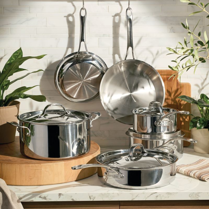 A lifestyle shot of the Anolon EverLast Tri-Ply Clad stainless steel 10-piece cookware set, featuring several pots and pans with lids arranged on a marble countertop and two frying pans hanging from a rack against a white tiled wall, accented by green plants and wooden cutting boards.