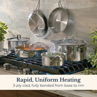A lifestyle shot of Anolon EverLast Tri-Ply Clad stainless steel cookware in use on a gas stovetop, with steam rising from a small saucepan of greens and a sauté pan of salmon. Two pans hang on a tiled wall in the background. Text reads: "Rapid, Uniform Heating. 3-ply clad, fully bonded from base to rim."