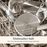 A close-up shot of Anolon EverLast Tri-Ply Clad stainless steel pots and pans arranged in a dishwasher rack, with the bottom of one pan clearly showing the Anolon logo. Overlay text reads: "Dishwasher Safe – For the demands of the busy kitchen."