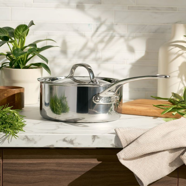 An EverLast Tri-Ply Clad Stainless Steel 3-Quart Saucepan with Lid rests on a marble counter, surrounded by green plants, a wooden cutting board, a light vase, and a beige towel—essential cookware for home chefs. White tile backsplash behind.