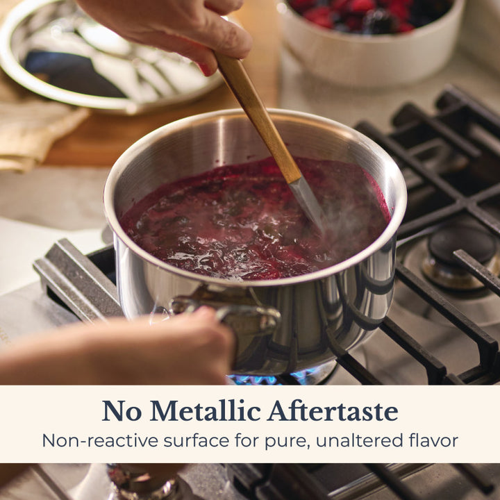 A person stirs a pot of dark purple liquid on a gas stove. Text overlay: No Metallic Aftertaste. The EverLast Tri-Ply Clad Stainless Steel 3-Quart Saucepan with Lid has a non-reactive surface for pure flavor. A bowl of berries is in the background.
