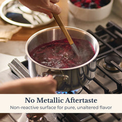 A person stirs a pot of dark purple liquid on a gas stove. Text overlay: No Metallic Aftertaste. The EverLast Tri-Ply Clad Stainless Steel 3-Quart Saucepan with Lid has a non-reactive surface for pure flavor. A bowl of berries is in the background.