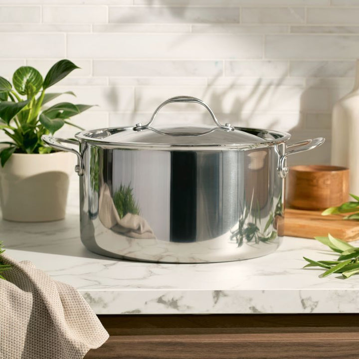 An EverLast Tri-Ply Clad Stainless Steel 8-Quart Stockpot with Lid sits on a marble kitchen countertop, surrounded by green potted plants and cookware accessories in a bright, modern setting.