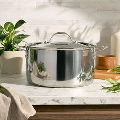 An EverLast Tri-Ply Clad Stainless Steel 8-Quart Stockpot with Lid sits on a marble kitchen countertop, surrounded by green potted plants and cookware accessories in a bright, modern setting.