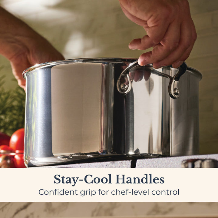 A person holds the handle of an EverLast Tri-Ply Clad Stainless Steel 8-Quart Stockpot with Lid on a stovetop. Text reads: Stay-Cool Handles. Confident grip for chef-level control. Tomatoes and herbs can be seen in the background.