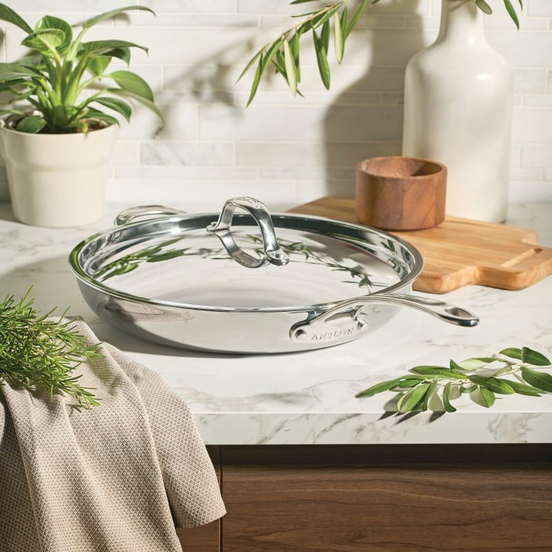 The EverLast Tri-Ply Clad Stainless Steel 12.5-Inch Frying Pan with Lid and Helper Handle rests on a marble countertop among kitchen accents—a must-have cookware essential for every home chef.