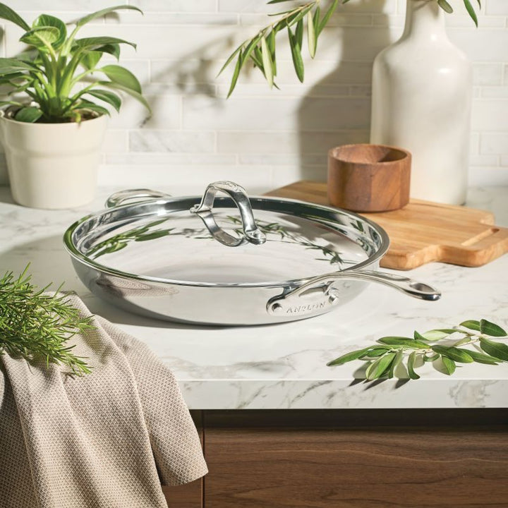 The EverLast Tri-Ply Clad Stainless Steel 12.5-Inch Frying Pan with Lid and Helper Handle rests on a marble countertop among kitchen accents—a must-have cookware essential for every home chef.