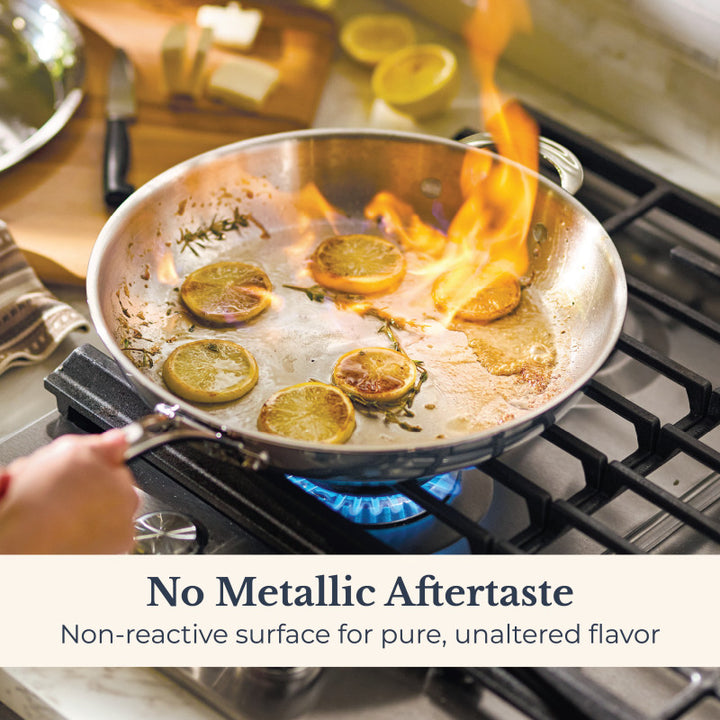 A home chef sears lemon slices in the EverLast Tri-Ply Clad Stainless Steel 12.5-Inch Frying Pan with Lid and Helper Handle over a gas stove, flames rising. Text below: No metallic aftertaste—non-reactive surface for pure flavor.