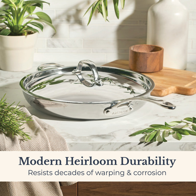 An EverLast Tri-Ply Clad Stainless Steel 12.5-Inch Frying Pan with Lid and Helper Handle rests on a marble countertop amid herbs, a wooden container, and a plant. Text reads: Modern Heirloom Durability—resists decades of warping & corrosion.