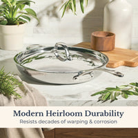 An EverLast Tri-Ply Clad Stainless Steel 12.5-Inch Frying Pan with Lid and Helper Handle rests on a marble countertop amid herbs, a wooden container, and a plant. Text reads: Modern Heirloom Durability—resists decades of warping & corrosion.